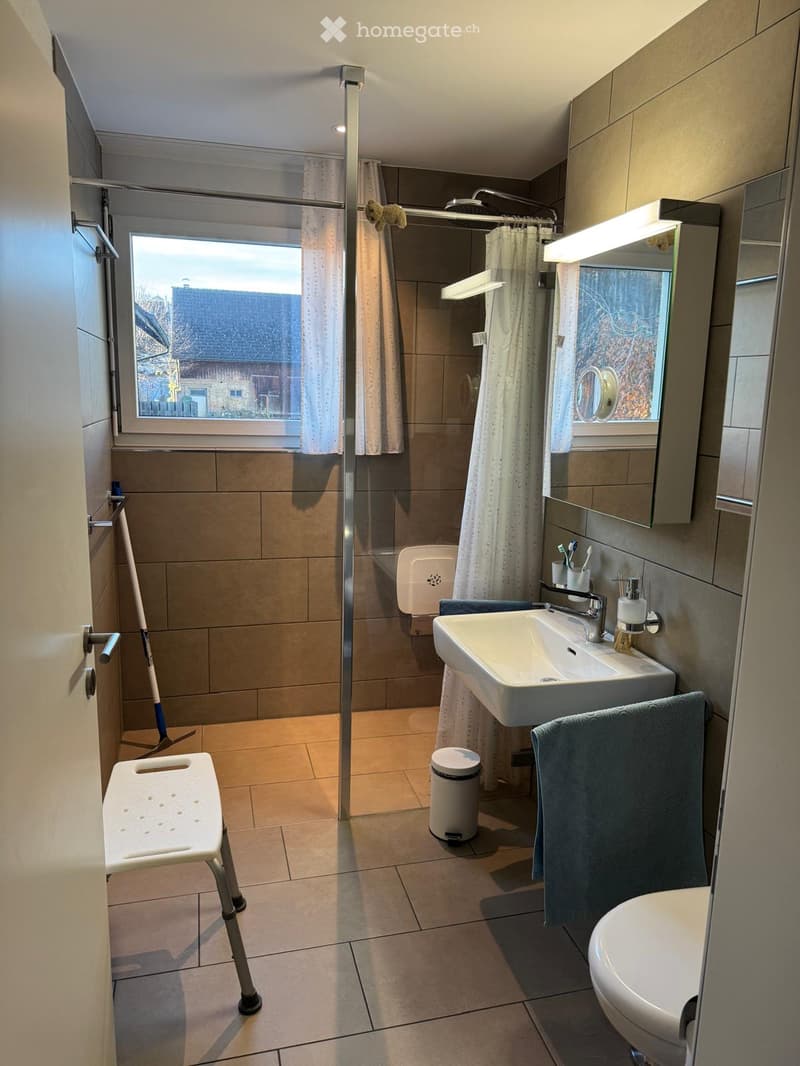 Modern bathroom with a shower, sink, toilet, mirror, and a towel on the floor. The room features tiled walls and floor, a window with a view, and a small stool near the door.