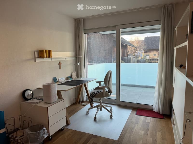 A modern, well-lit home office with a wooden floor, large window, and sliding glass door leading to a balcony. The room features a desk with a chair, a lamp, a clock, and a shelf with books.