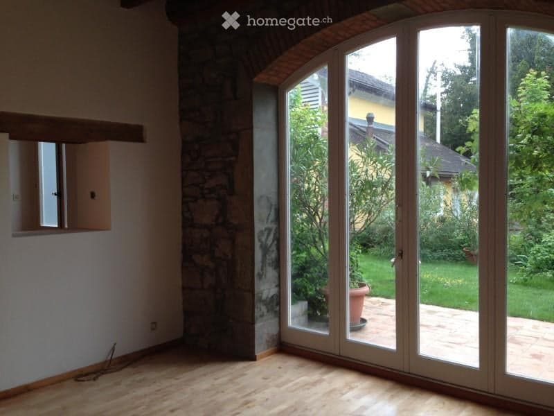 Empty room with wooden flooring, large glass sliding doors leading to a garden, and a stone wall on one side.
