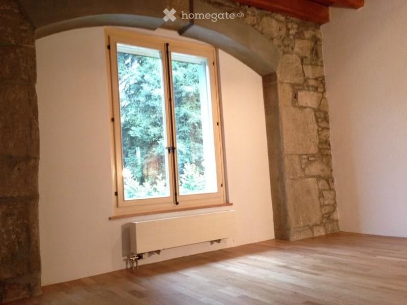 Empty room with wooden flooring, white walls, a large window with wooden frame, and a radiator below the window. The room has a stone wall on one side and an arched ceiling.