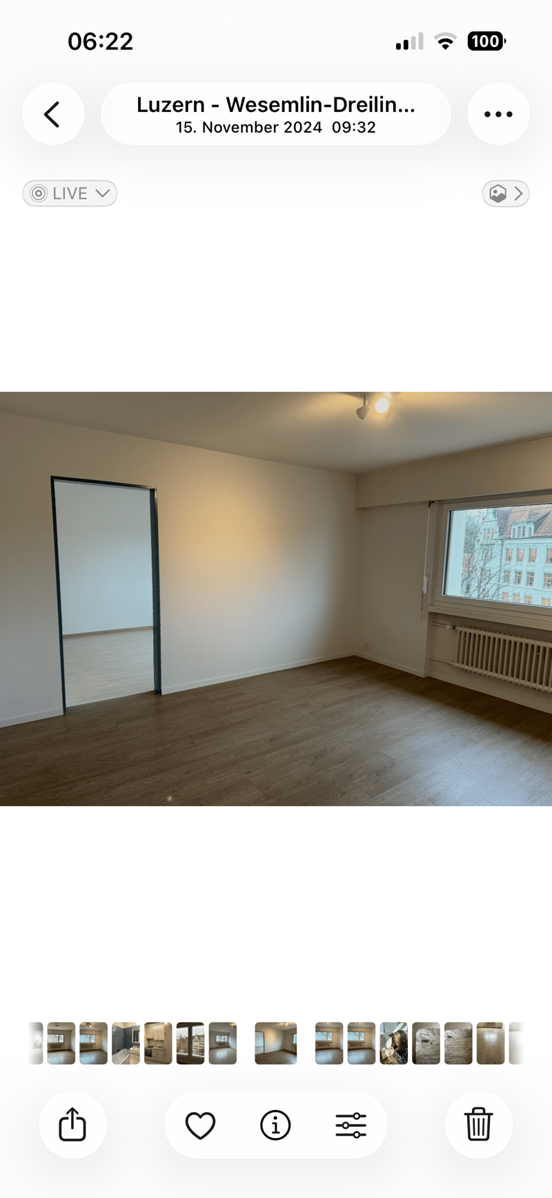 Empty room with wooden flooring, white walls, and a large window with a view of a building outside. The room has a door on the left side and a radiator below the window on the right.