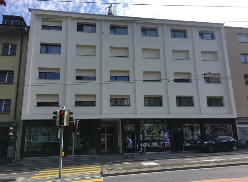 The facade of a white building with many windows and an entrance, traffic lights on the left and a car parked in front on the right.