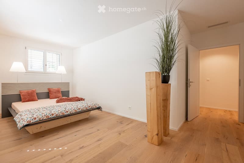Modern bedroom with wooden flooring, a bed with a colorful bedspread and pillows, two table lamps, and a tall potted plant near the door.