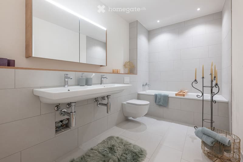 Modern bathroom with double sink, toilet, bathtub, large mirror, decorative candles, and a furry rug on the floor.