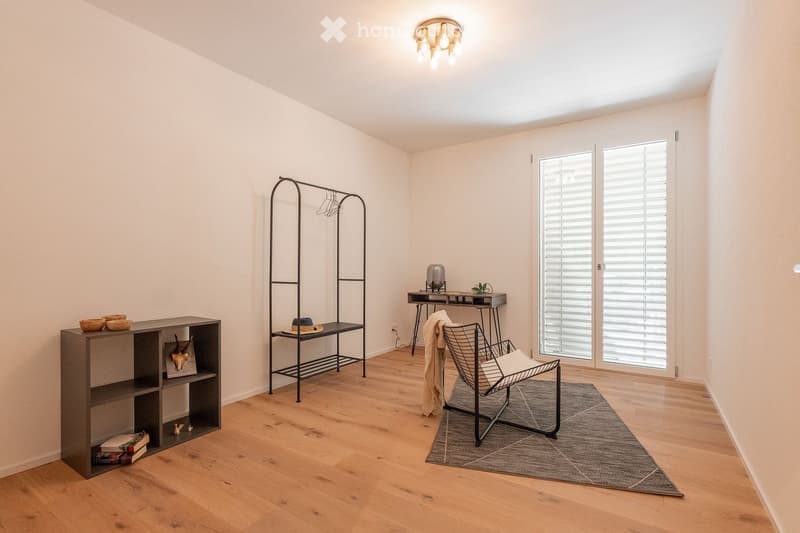 A modern, minimalist room with light wooden flooring, white walls, and large windows with blinds. Features include a sleek black metal chair, a small table with a lamp, a shelf with decorative items, and a gray rug.