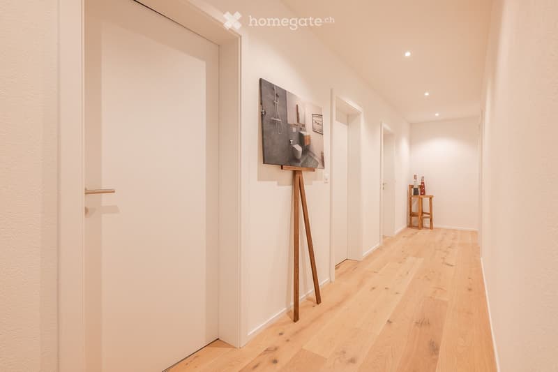 A modern, minimalist hallway with light wooden flooring, white walls, and recessed lighting. Features include a large painting on an easel, a small wooden table with decorative items, and a partially visible bathroom with modern fixtures.