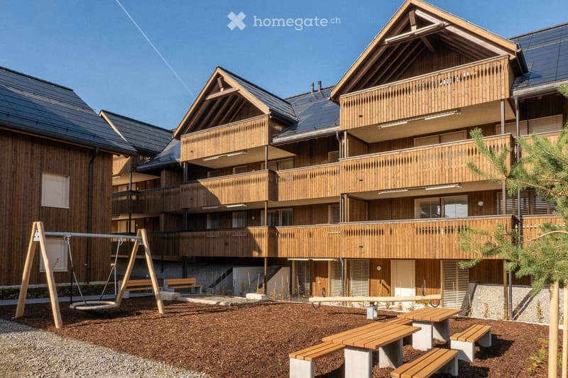 A multi-story wooden building with balconies and a playground area featuring benches and a swing set.