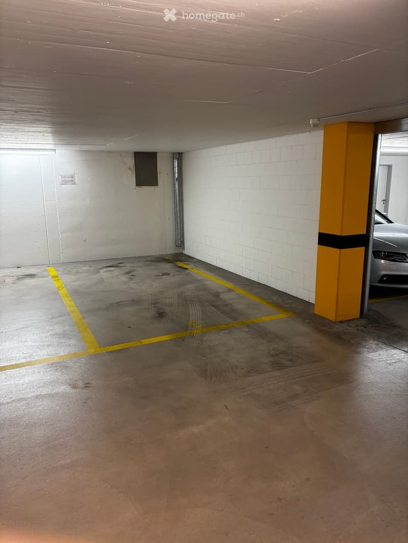 Empty underground parking space with concrete floor, white brick walls, yellow parking lines, and a single yellow support pillar.