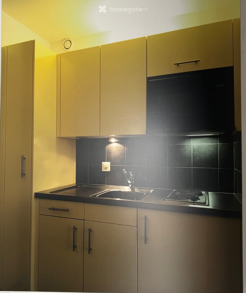 Modern kitchen with light-colored cabinets, a black countertop, a sink, a faucet, and a built-in stove. The backsplash is dark tiles, and the lighting is warm and focused.