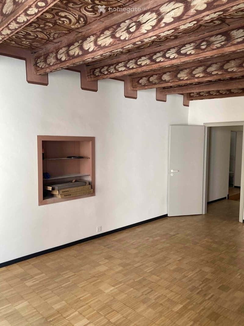Empty room with wooden flooring, white walls, and a decorative wooden ceiling. A built-in shelf holds a few boxes and items, and a door leads to another space.