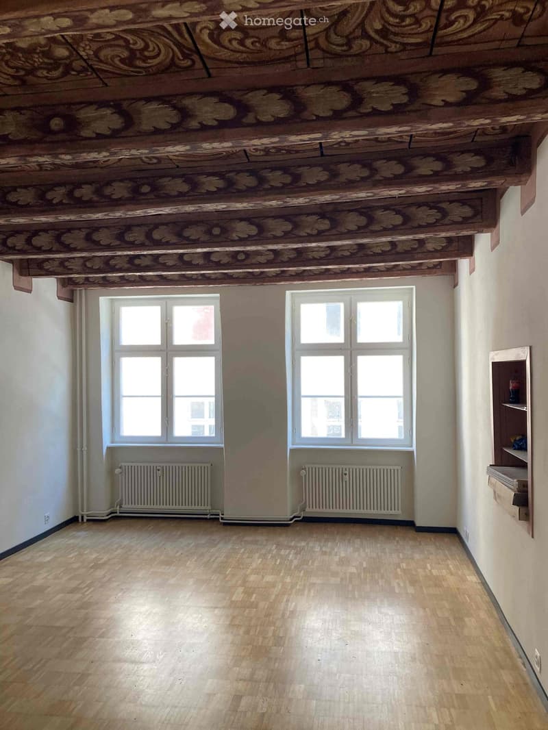 Empty room with wooden floor, exposed wooden ceiling beams, two windows, radiators, and a small shelf on the right side.