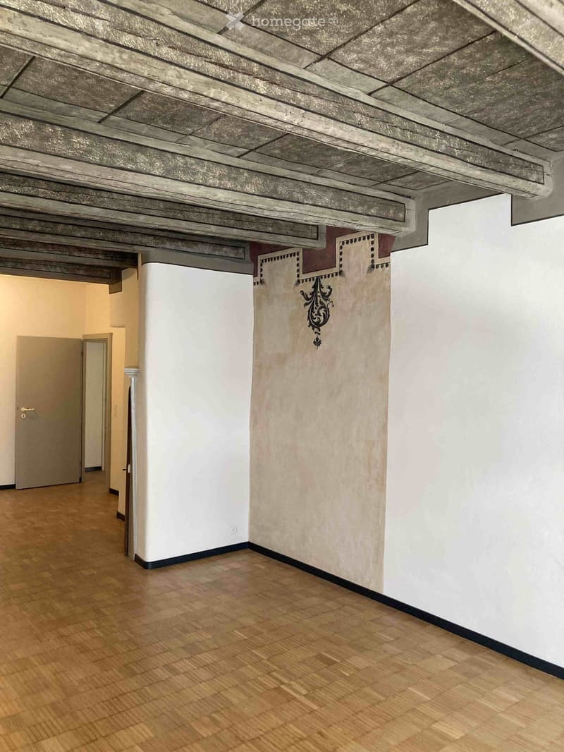 Empty hallway with wooden flooring, white walls, and exposed wooden ceiling beams. A decorative wall design is visible on one wall.