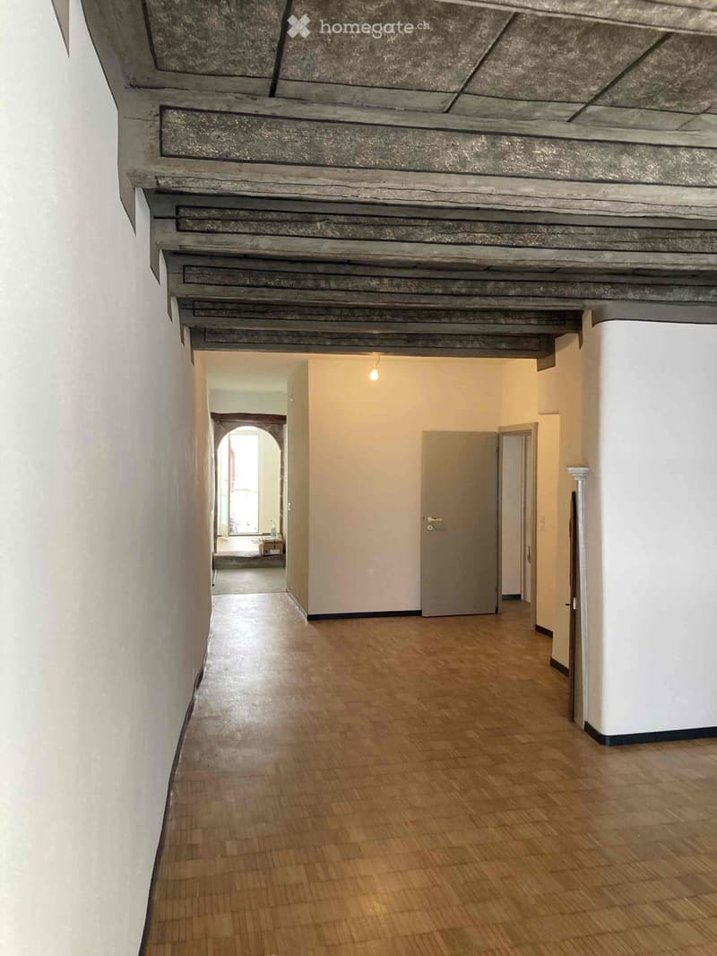 Wooden-floored hallway with white walls, arched doorway leading to another room, and visible ceiling beams.