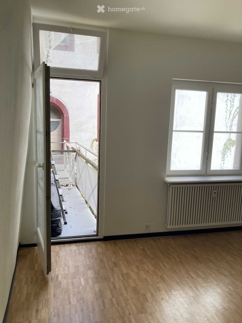 Empty room with wooden flooring, large window, radiator, and open sliding door leading to a balcony.