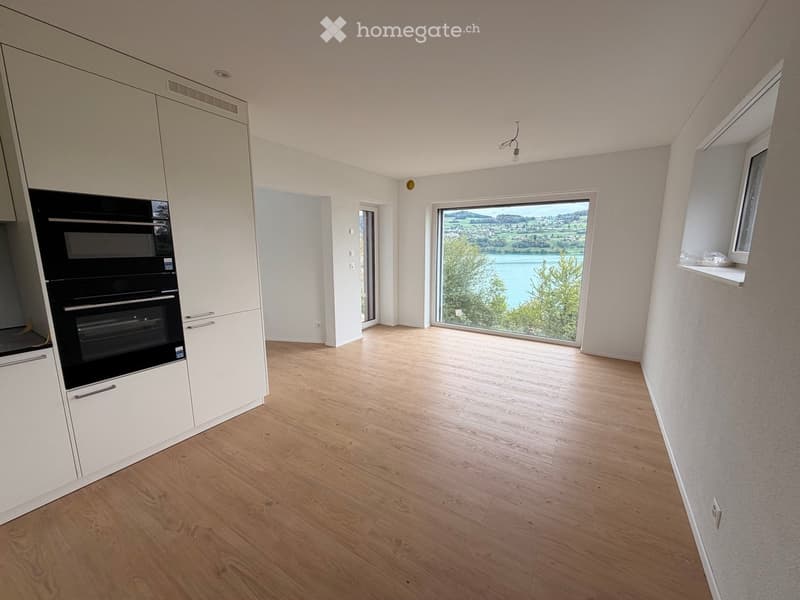 Empty room with light wooden flooring, white walls, and large windows offering a scenic view of a lake and surrounding landscape.