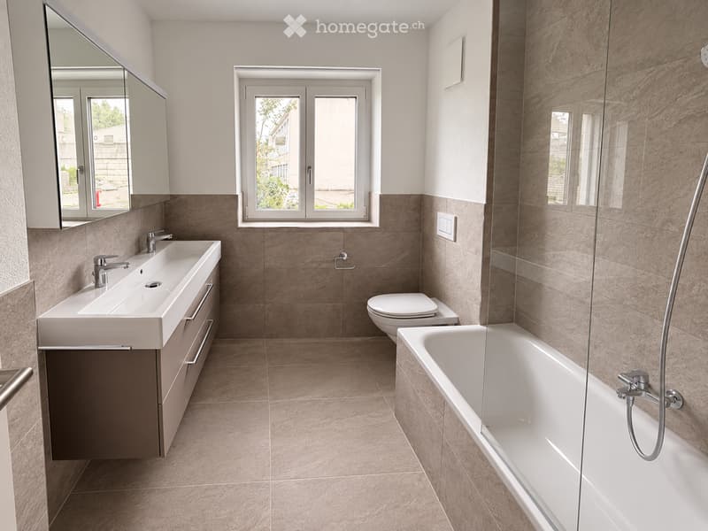 Modern bathroom with a double sink, toilet, bathtub, and large mirror. The space features beige tiles and a large window providing natural light.