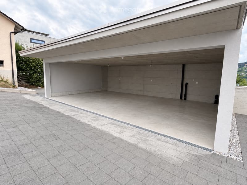 Modern garage with a concrete floor and walls, covered by a flat roof with exposed wooden beams. The entrance is a large, clean, grey garage door.