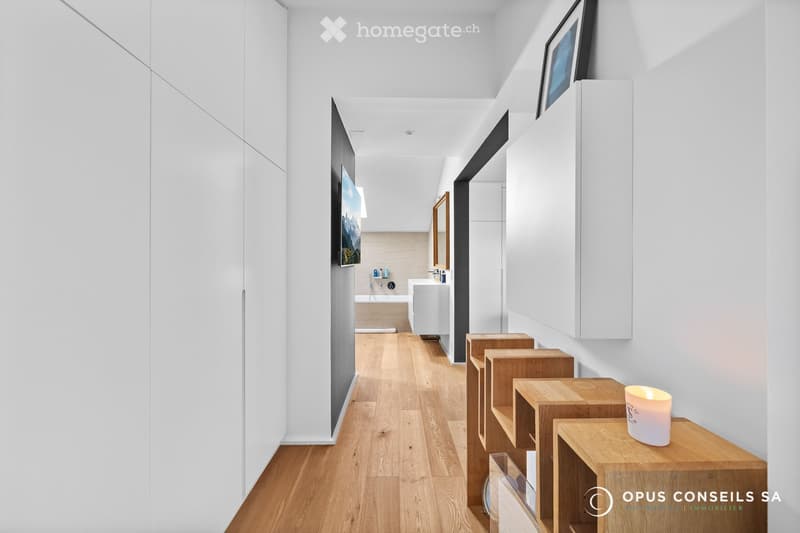 Modern hallway with light wooden flooring, white walls, built-in white cabinets, and a small candle on a wooden shelf. A bathroom is visible at the end of the hallway.