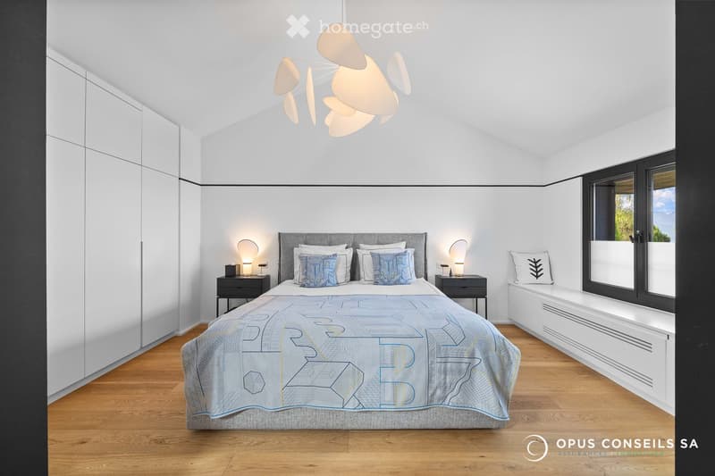 Modern bedroom with a large bed, nightstands, lamps, and a window with a view. The room features a contemporary design with white walls, wooden flooring, and built-in storage.