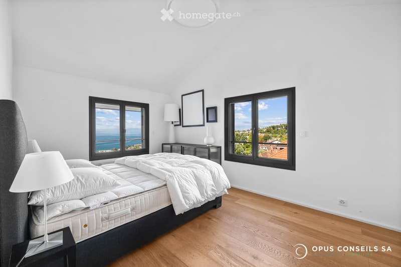 Bright bedroom with large windows offering scenic views, featuring a neatly made bed, nightstand, lamp, dresser, and decorative mirrors.