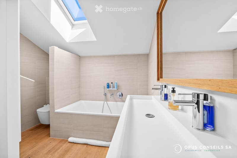 Modern bathroom with a bathtub, toilet, sink, and mirror. Features a skylight window and wooden flooring.