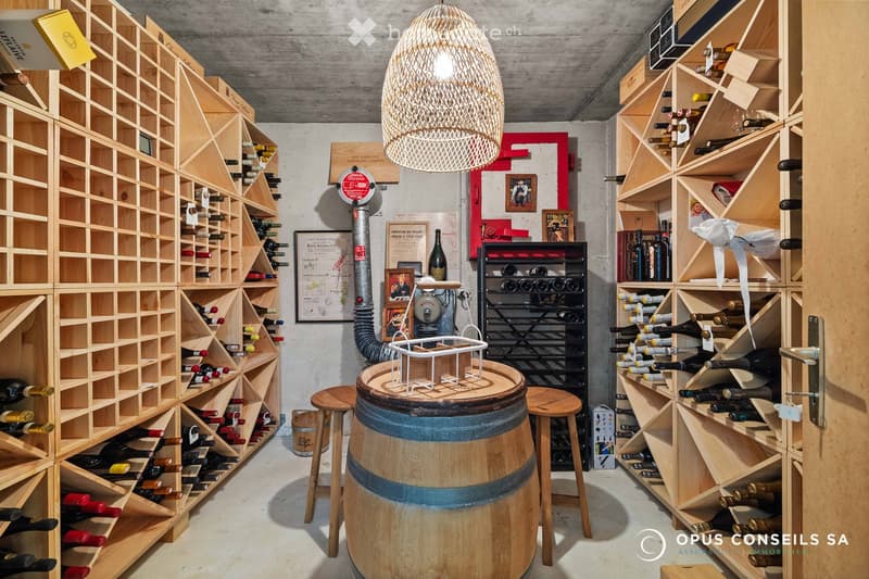 A wine cellar with wooden racks filled with bottles, a large wine barrel in the center, a hanging pendant light, and framed pictures on the walls.