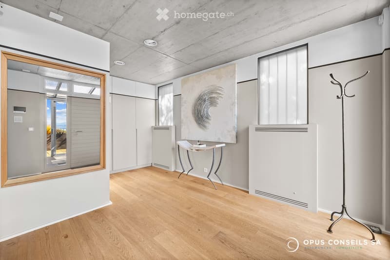 Modern, minimalist room with light wooden flooring, white walls, and contemporary decor including a small table, chair, coat rack, and a large mirror. The room features built-in storage units and a radiator.