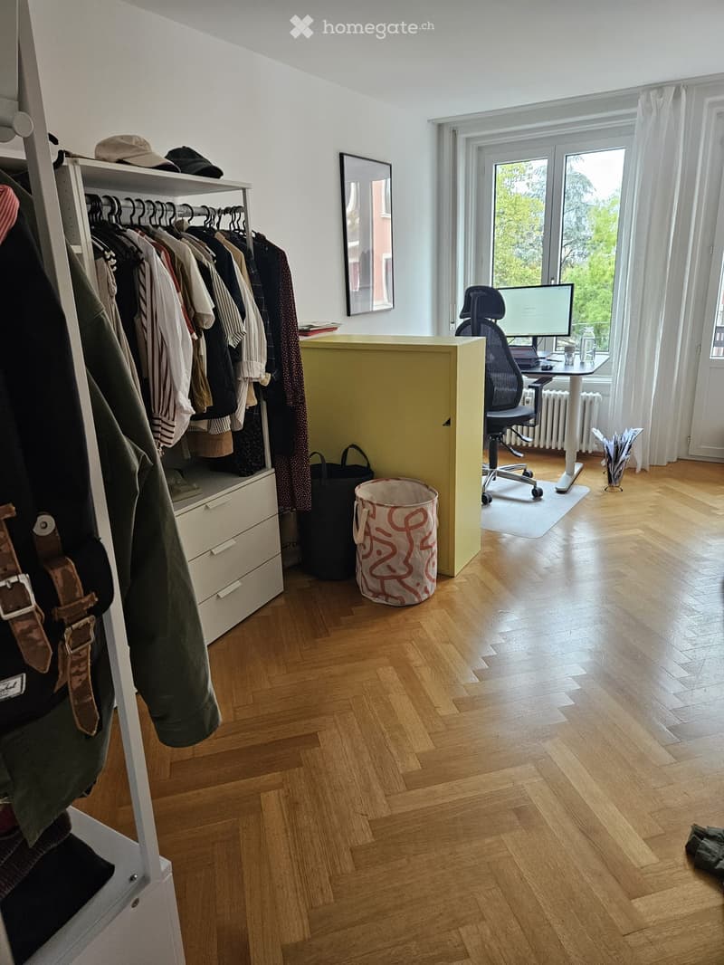 A well-lit room with wooden flooring, a wardrobe with hanging clothes, a desk with a chair, a laptop, and a mirror on the wall.