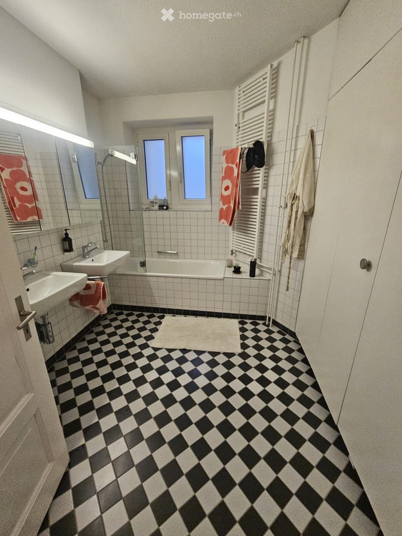 Bathroom with black and white checkered floor, two sinks, bathtub, mirror, towel rack, towels, and a small window.