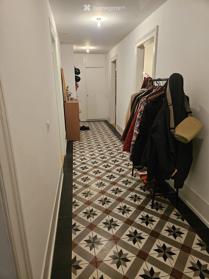 Hallway with patterned tile floor, coat rack with jackets, small cabinet, and a plant on the left side.