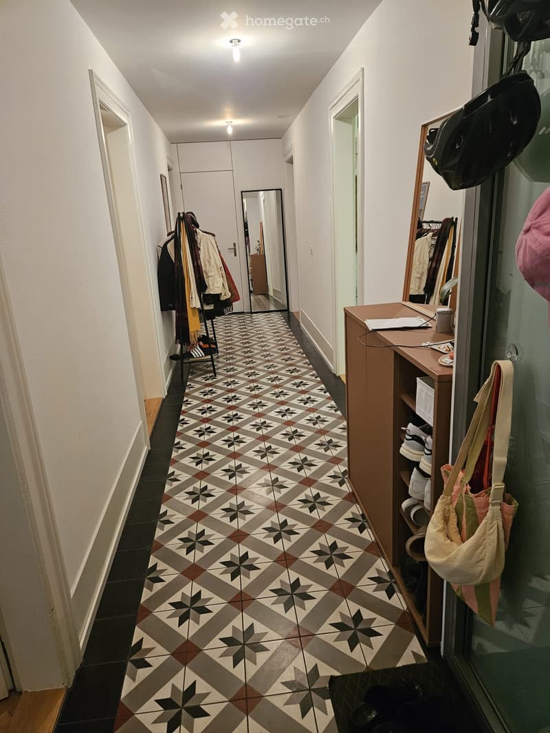 Hallway with patterned tile floor, white walls, and several doors leading to other rooms. A coat rack with jackets and a small table with a bag and shoes are visible.
