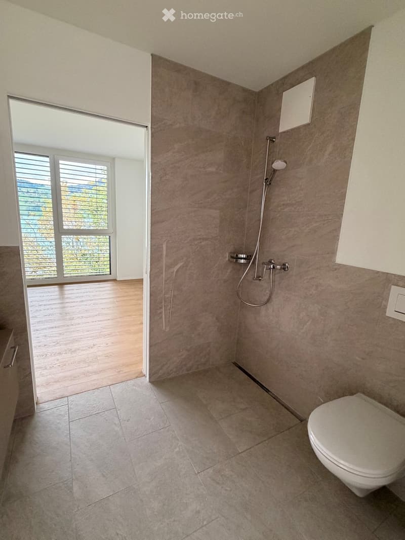 Modern bathroom with tiled walls and floor, a toilet, and a shower with a handheld sprayer. The room features large windows with blinds, allowing natural light to enter.