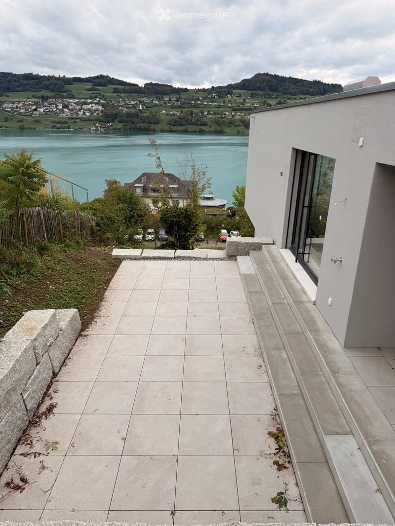 Modern concrete balcony with steps leading up, overlooking a scenic lake and town with distant hills.
