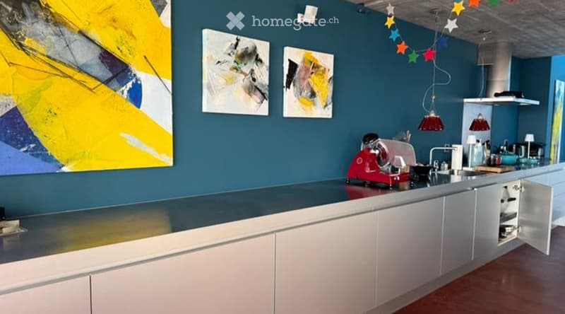 Modern kitchen with white cabinets, dark blue walls, and a sleek black countertop. Features a red appliance, pendant lights, and colorful artwork.