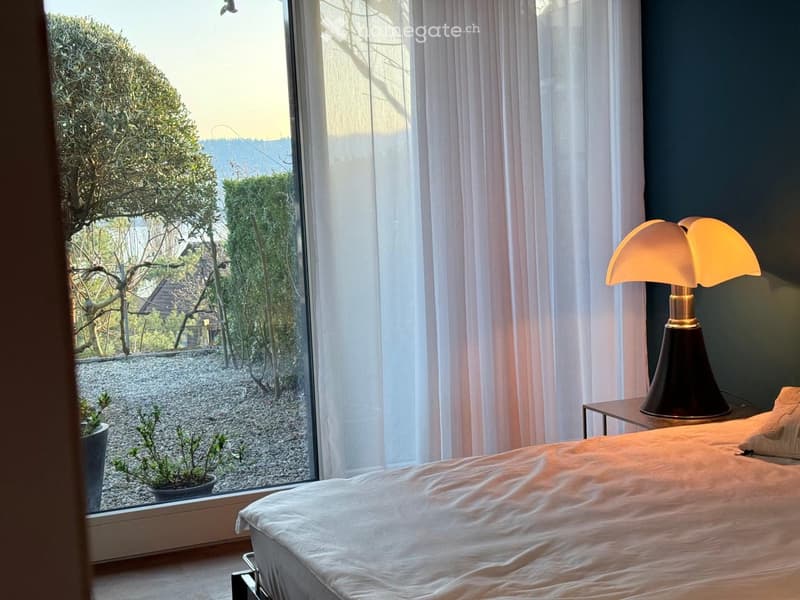 Modern bedroom with a large window offering a view of a garden, featuring a neatly made bed, a table lamp on a nightstand, and a small potted plant on the windowsill.