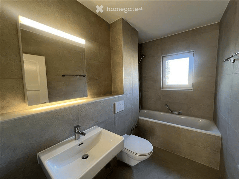 Modern bathroom with a sink, toilet, bathtub, and a large mirror with built-in lighting. The walls are tiled in neutral tones, and the space is well-lit with natural light from a small window.