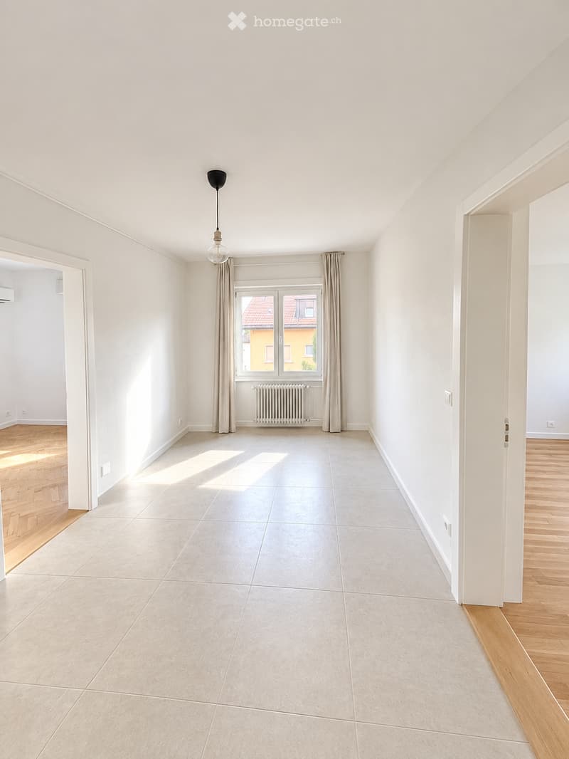 Empty room with white walls, light wooden flooring, and a large window with curtains. The space is clean and unfurnished, with a radiator below the window and a pendant light hanging from the ceiling.