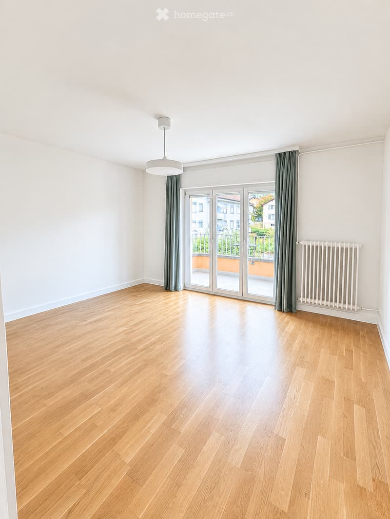 Empty room with light wooden flooring, white walls, large sliding glass doors leading to a balcony, and a radiator on one wall.