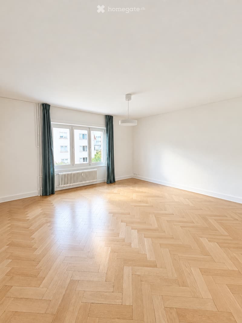 Empty room with light wooden herringbone flooring, white walls, large window with dark curtains, radiator, and a single ceiling light fixture.