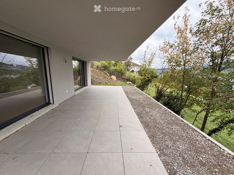 Modern balcony with large glass windows, concrete tiles, and a view of greenery and distant landscape.