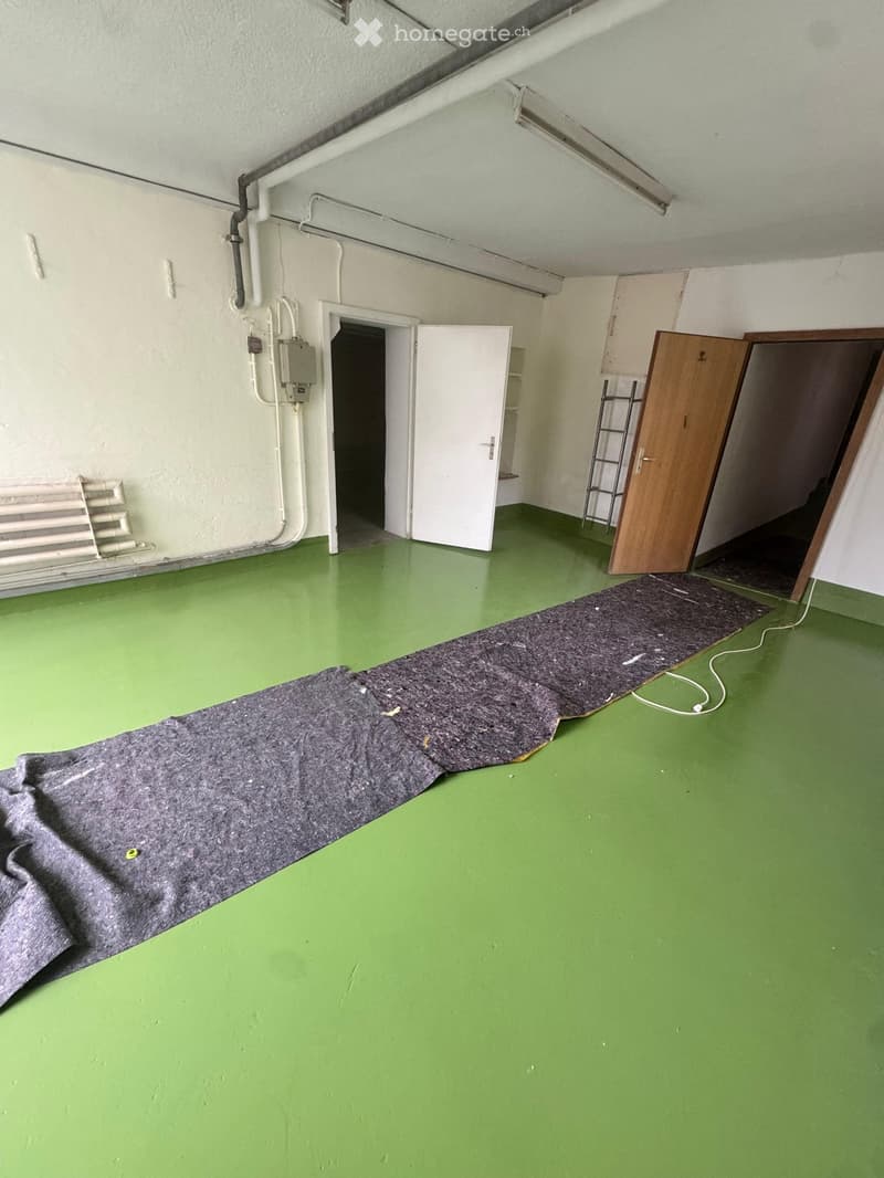 Empty room with green floor, white walls, two doors, one closed, one open, a ladder and a radiator on the wall