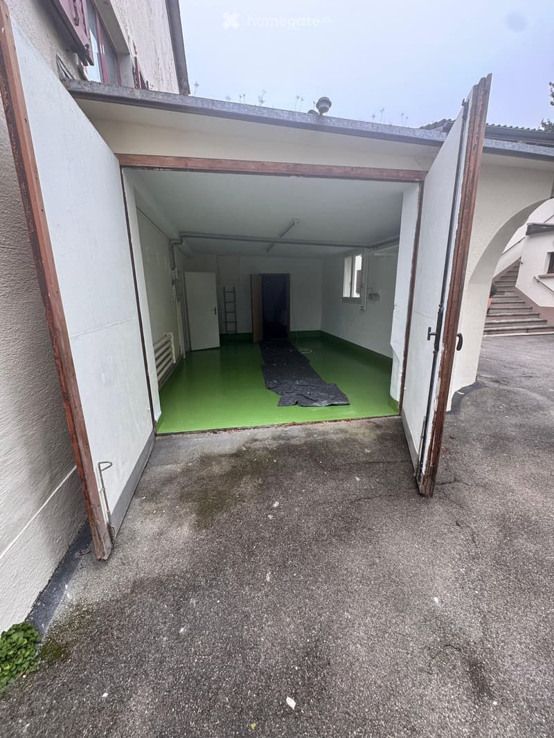 A closed garage with green floor, an opened door, a ladder, and two windows.
