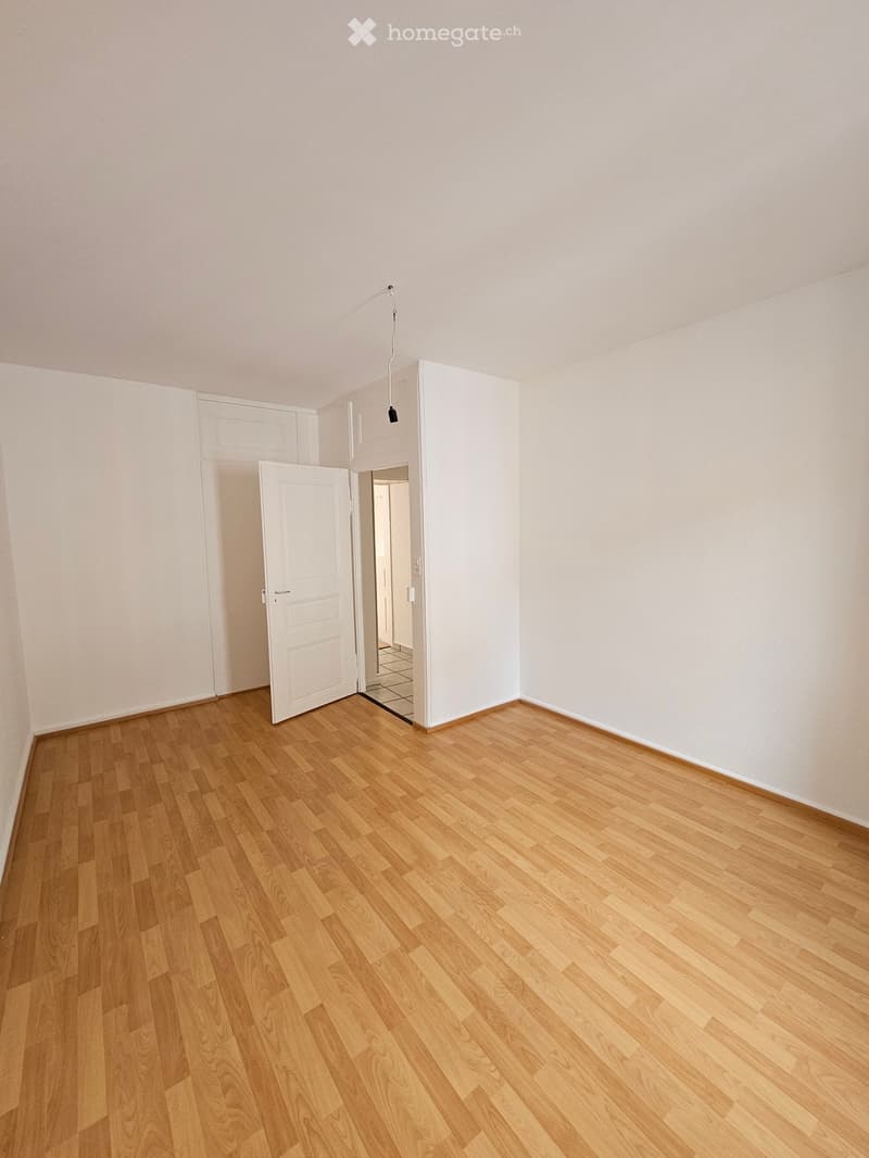 Empty room with light wooden flooring and white walls, featuring an open doorway leading to another space.