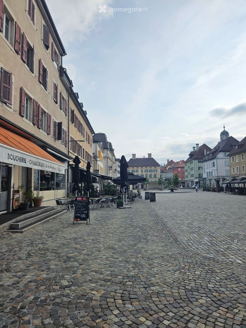 A cobblestone square with outdoor seating, closed umbrellas, and buildings with shutters and awnings.