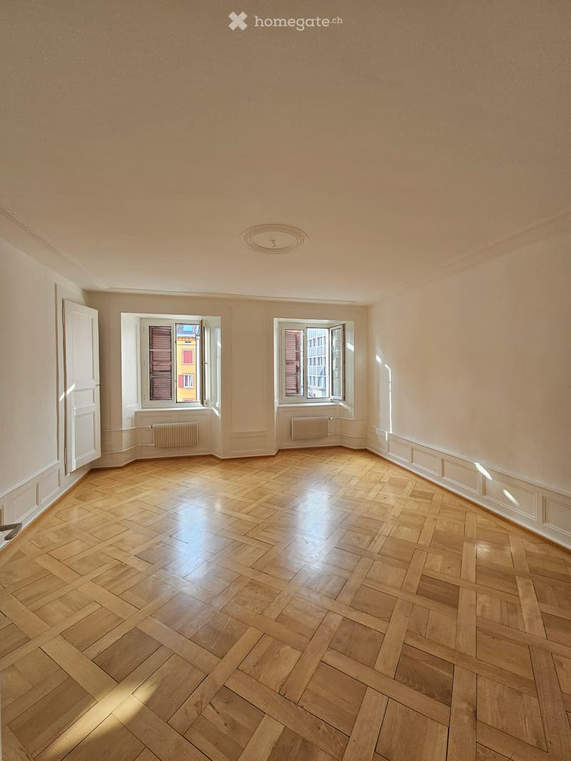 Empty room with light wooden herringbone flooring, white walls, and two large windows with open shutters allowing natural light to enter.