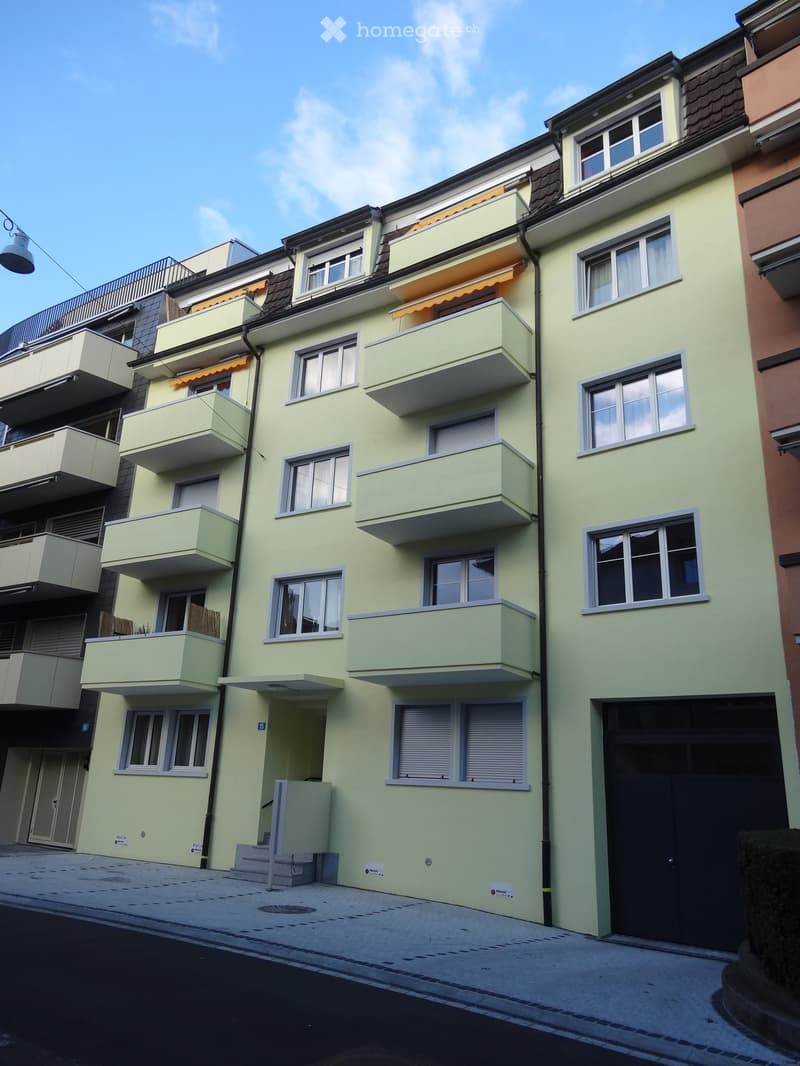 Four-story residential building with light green facade, multiple windows, balconies with awnings, and a garage entrance.