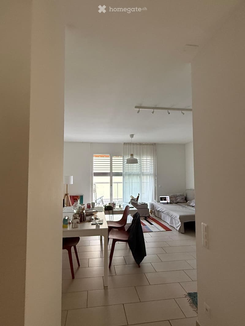 A modern, well-lit room with a bed, a table with chairs, a laptop, and a curtained window. The space features tiled flooring and a colorful rug near the bed.