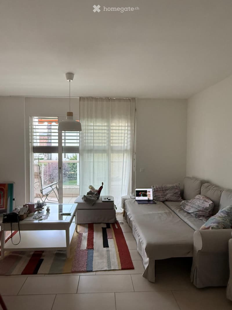 Living room with a large window and balcony access, featuring a sectional sofa, coffee table, laptop, and pendant light.