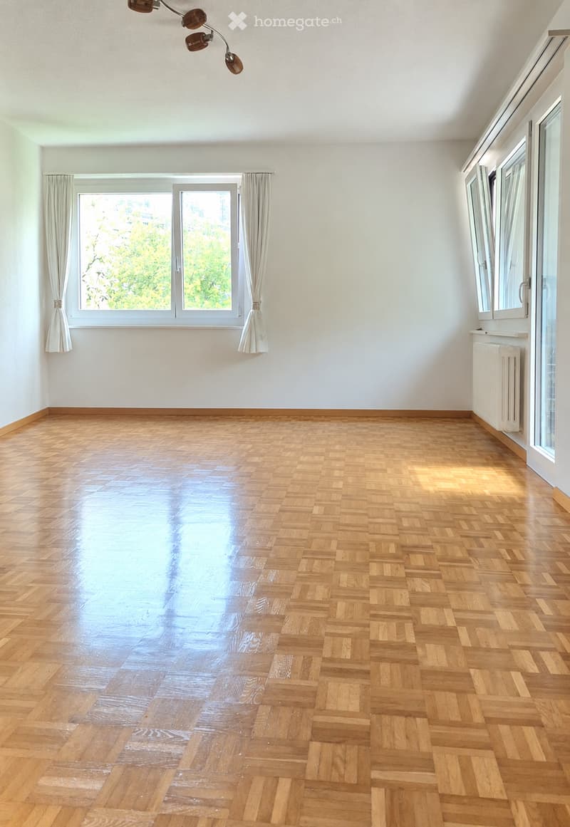 Empty room with wooden parquet flooring, white walls, large windows with curtains, and a radiator on the right side.