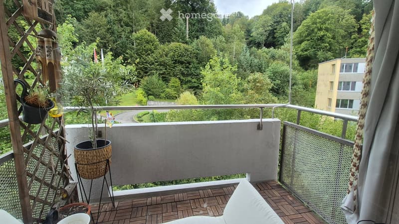 Balcony with wooden decking, metal railing, potted plants, and a view of a green, wooded area with a building in the distance.